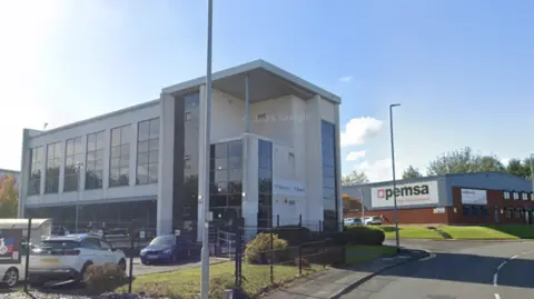 A modern three-storey office building with large glass windows and white exterior cladding stands beside a road on a sunny day. Several cars are parked in front of the building behind a black fence, and a lamppost is visible in the foreground. To the right, a neighbouring commercial building displays the sign “Opemsa Management.” The main office building has signage near the entrance, though the text is not clearly legible. The sky is bright blue with a few small clouds, and landscaped grass and shrubs surround the properties.