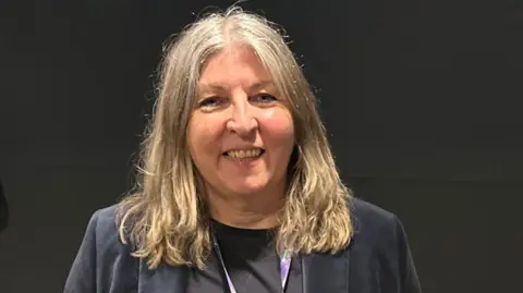 Chrissy Callaghan, who has shoulder-length light brown hair, is wearing a dark blue jacket over a black top.