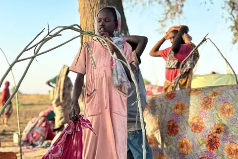 AFP via Getty Images A Sudanese girl in an orange dress starts to build a shelter from sticks and pieces of material after fleeing el-Fasher.
