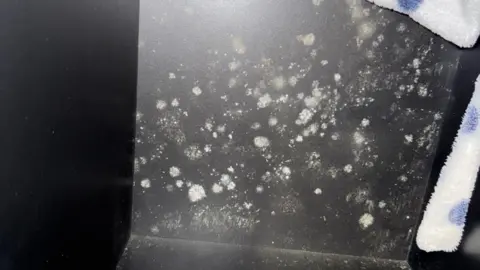 Jessica White and light green mould spores on a black wardrobe. The robe of a dressing gown can be seen hanging in the corner, it is white with blue spots.