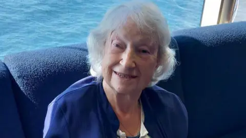 Wendy Hill A woman with short white hair layered around her face, smiling while sat inside on a boat. Blue sea can be seen in the background and she is on a cushioned navy seat. She is wearing a navy cardigan and striped white and blue top.