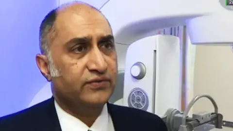 Amir Bhatti wearing a dark suit jacket, white shirt, and red striped tie standing indoors next to a large medical imaging machine with a control panel and arm mechanism. The background includes a softly lit wall and part of the machine’s structure.