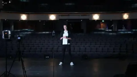 William Laven Young man standing on a stage in a dark theatre with rows of seats behind him