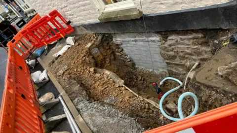 Olivia Richwald/BBC A hole in the pavement next to an old stone building painted white. The hole has water in the bottom of it and there is a hose and drainage kit in there. It is surrounded by red plastic barriers.