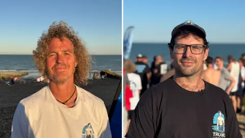 Both smile into the camera and are standing on the beach. Mark Trigg has curly hair and is wearing a white T-Shirt. Ben Windsor has a cap and T-Shirt on both branded Trunks and Hunks
