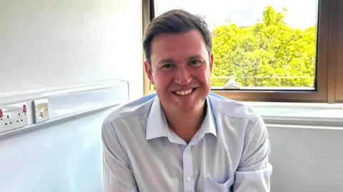 Steve Race MP Steve Race. He has short brown hair and is smiling at the camera. He is wearing alight blue shirt. He is sat in an office with a window behind him. 