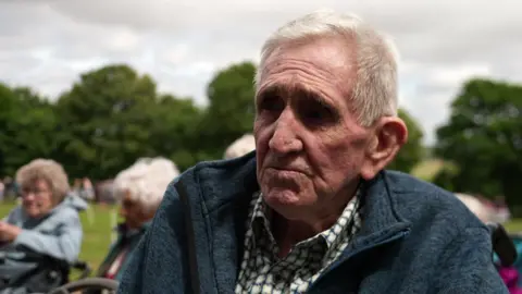 An older man with short white hair wears a green and white checked shirt and a blue jacket.