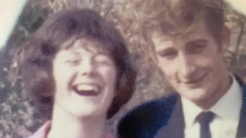 Mark Payne A blurry photo of a young Mark Payne with his mum, Gill Payne. Gill is laughing and has short brown hair. Mark is smiling at the camera wearing a dark suit and has brown wavy hair.