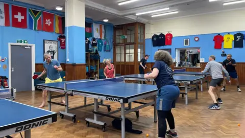 Several people are playing table tennis across multiple tables in an indoor hall with blue walls. Flags and club T‑shirts hang on the walls and orange balls are on the floor.