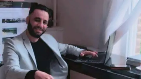 Adam Mejri, a man with a short dark beard and short dark hair, sits a desk turned towards the camera, smiling. He wears a light grey blazer and black top.