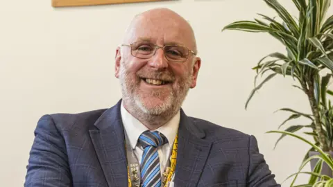 Paul Norton A man with a beard and gold-rimmed glasses wearing a blue checked jacket, a striped tie and lanyard
