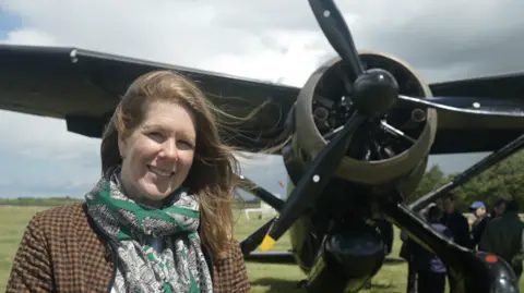 Jamie Niblock/BBC A woman with long brown hair smiles at the camera as she stands in front of an old style war propeller aircraft. She wears a chequered brown and black jacket and a large scarf with green and white patterns on it.