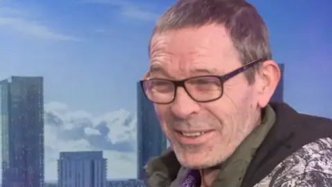 Greg Mounfield has short greying dark hair, dark glasses, khaki cardigan, and a hoodie with a black lining white, grey and black paisley pattern.