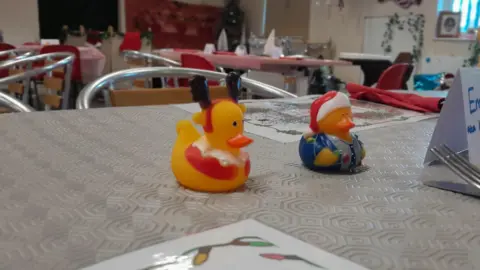 A close-up of two Christmas themed rubber ducks on a table. In the background there are more tables, which are adorned with tablecloths and decorations.