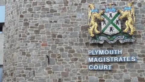 Outside of Plymouth Magistrates' Court. There is a beige stone wall and a crest. Underneath the crest is writing that says Plymouth Magistrates' Court.