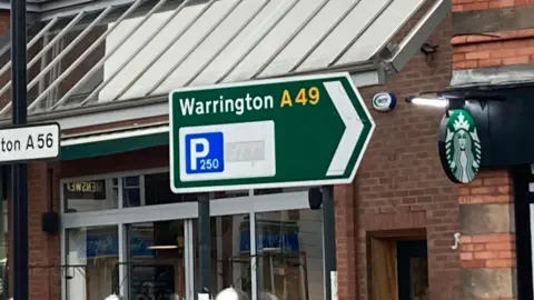 Image of a green road sign that reads Warrington A49, which is pointing right and has a blue square with a P for parking in it. The 'free' part of the sign has been blocked out 