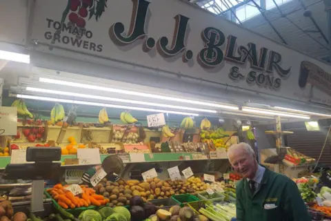 Victoria Clark Robin Blair is standing in front of the colourful fruit and vegetables at his stall and smiling. He has short white hair and is wearing a traditional green jacket. The green and white sign above him reads J.J Blair & Sons.