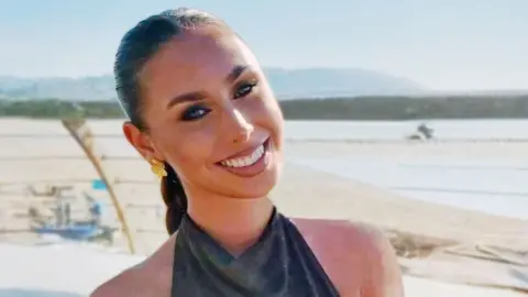 Family handout Erin has brown hair tied back and smiles at the camera. There is a beach behind her