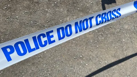 BBC A picture of blue and white police tape. The tape itself is white with the writing 'POLICE DO NOT CROSS' along it in blue text.