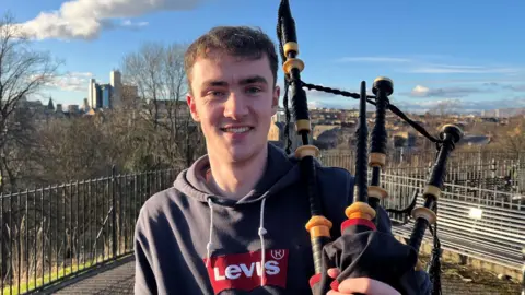 BBC Alasdair has brown hair and is smiling. He is resting the pipes on a shoulder. Behind him is a metal fence and a Glasgow skyline. 