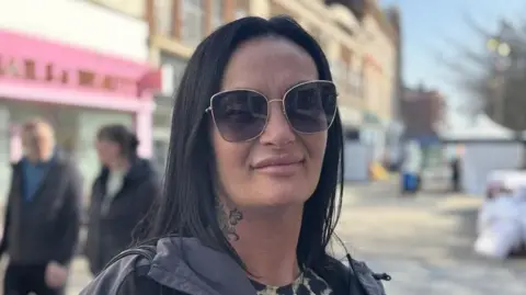 Kimberly poses in the high street. She has drak hiar and wears dark shades and a jacket.