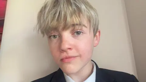 Ben Ben has green eyes and blonde hair that comes down over his eyebrows. He is wearing a dark blazer and white shirt. 