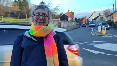 Susannah Clemence Susannah Clemence is standing by a road with a mini roundabout with cars going by behind her. She is wearing a nacy coat and a colourful scarf and is smiling at the camera. 