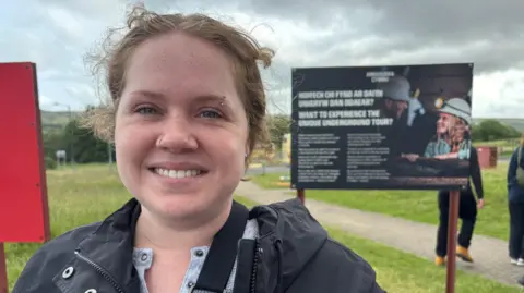 Woman dressed in black coat looking at the camera with big pit underground tour sign in the background.