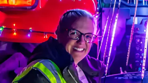 Andrew Mutimer Liz Quickenden wearing glasses and a high-vis vest over a dark coloured jacket. She is smiling and looking into the camera while standing in front of some colourful lights.