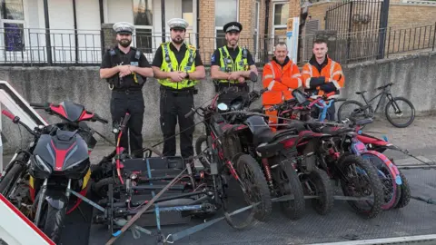 Dorset Police A number of e-bikes and motorbikes are strapped to a trailer. There are three police officers in uniform stood behind and two men wearing a florescent orange uniform. They are outside standing next to some black railings in front of buildings.