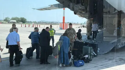SWNS Taken from behind, a group of people stand at the back of a plane which has a large ramp open. They have luggage, and soldiers help them on board.