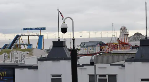 Tendring District Council A CCTV camera next to Clacton-on-Sea Pier. The pier is behind it. Off-shore wind turbines can be seen in the distance. 