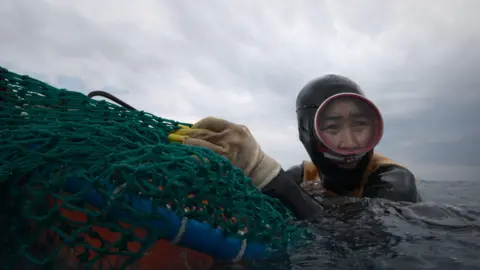 Apple  A haenyeo diver from South Korea’s Jeju Island in The Last of the Sea Women, wearing protective yellow gloves and diving gear as she holds on to a green fishing net