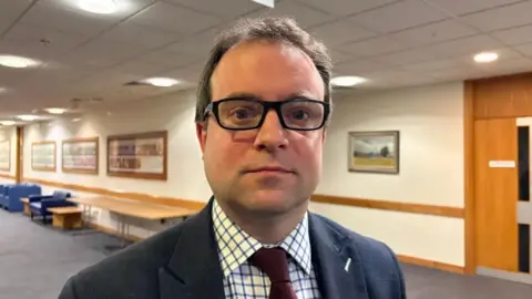 Martin Heath/BBC Close-up of Henry Vann wearing glasses, a dark suit jacket, a checked shirt and a red tie, standing in a brightly lit indoor hallway. Framed pictures line the walls behind, with tables and chairs along one side and a wooden door visible in the background.