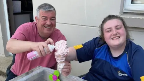 Conor and Ruby smiling at the camera. Conor has grey hair and is wearing a red t-shirt. Ruby has brown hair and is wearing a blue t-shirt.