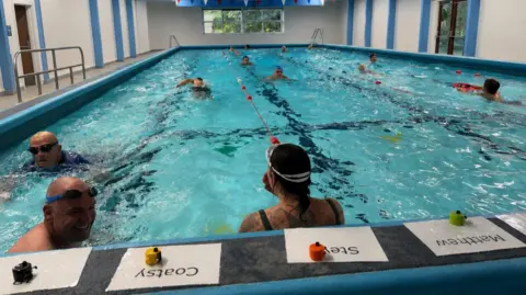 Laura Alliston The fundraising swim when it was ongoing. In relay style, one swimmer waits in the water whilst the other swims their lengths. The water is crystal-clear, chlorinated blue, which ripples as the swimmers power through. Some wear goggles.