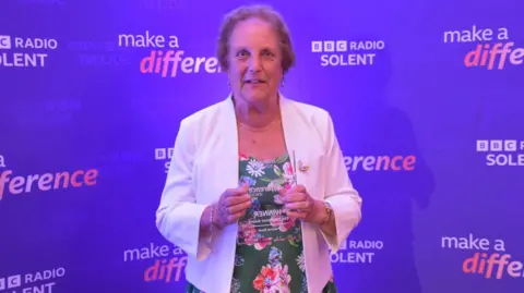 Pauline Steel holding her award, wearing a floral dress and a white jacket, in front of a purple wall, reading "BBC Radio Solent make a difference"