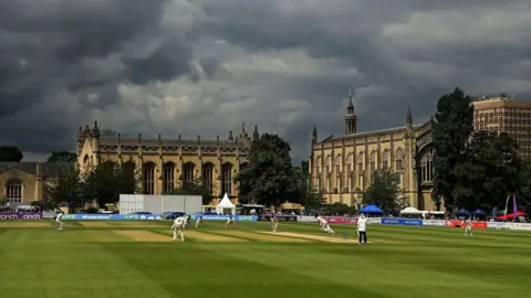 A wide view of cricketers from Gloucestershire and Hampshire playing during the Cheltenham Cricket Festival, with historic buildings in the background. The clouds are dark overhead