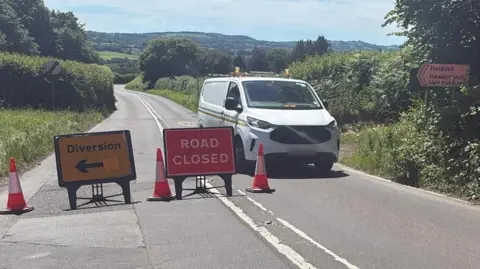 A white van parked on the road near the scene of a crash in the countryside. Road closed and diversion signs are on the road along with three traffic cones.