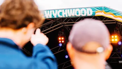 Wychwood Festival Two men point at the stage in the distance with 'Wychwood' visibile at the top in big white letters