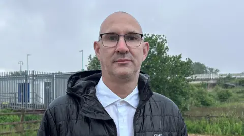 Kate Bradbrook/BBC Mick Lancaster, in a black coat with a hood and a white buttoned shirt. He is wearing eye glasses and standing in a car park near fencing.