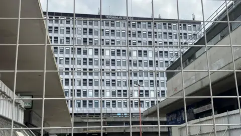 Terminus House in Harlow, seen through a large metal fence. It is an imposing silver building, formerly an office block, with lots of windows spread across it.