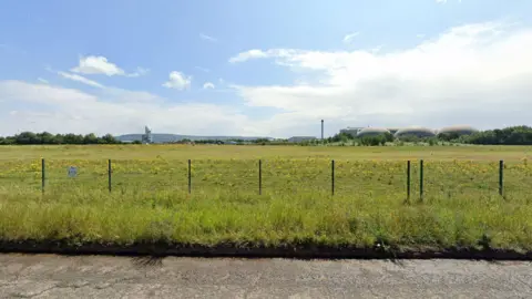 A grassy field with some industrial buildings in the distance.