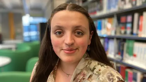 Verity Verhoven is looking straight into camera and in the background you can see a library area at the University of Derby. 
