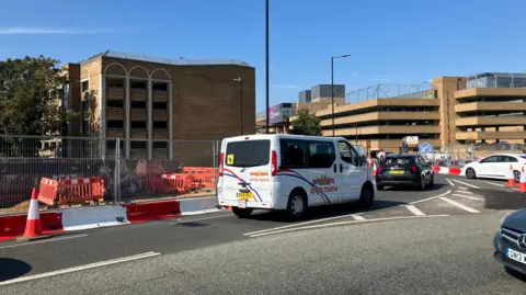 Shariqua Ahmed/BBC Cars driving along a roundabout with fencing on the left side of the road.