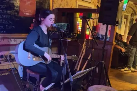 Stevie, a teenage girl with tied-back , dark hair, is sitting on a bar stool playing a blue acoustic/electric guitar and singing into a mic.