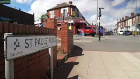 BBC The road sign is up close in the image with a street scene n the distance - a car is parked by some shops with houses on the opposite side of the road.