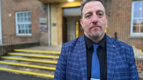 Phil Matthews-Dawson outside Devizes Police Headquarters. He is wearing a checked blue jacket, a dark shirt and a bright blue tie. He has a goatee beard and dark hair