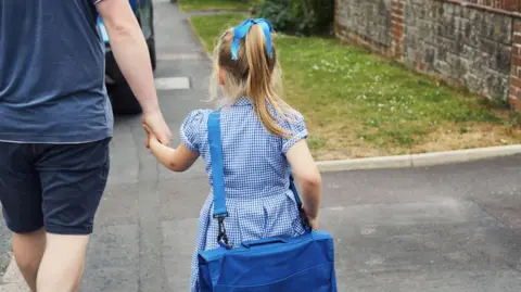 A young girl is seen walking down a pavement from behind. She is wearing a blue dress and is carrying a book bag. She is holding a parent's hand.