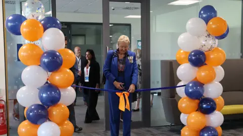 HUC Julie Spence cutting a blue ribbon at a doorway that is surrounded by a multicoloured balloon arch.